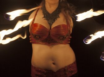 This photo features the leather costume for the fire artist more closely. There are carvings and painted flowers visible on the top, also the armour-like scaled style is more noticeable. Artist holds sets of burning torches on both sides on the photo edges.