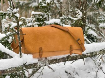 Very large leather bag is photographed in the snowy forest. Bag is set onto the fallen tree branch and is surrounded by snowy spruce branches.