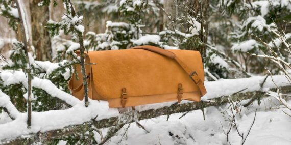Very large leather bag is photographed in the snowy forest. Bag is set onto the fallen tree branch and is surrounded by snowy spruce branches.