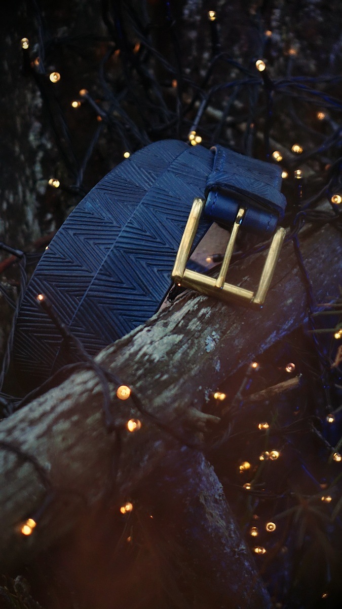Photo has been taken in the darkening forest scenery. Belt is placed onto the tree branch, there is a tree trunk visible on the background and some sparkling little lights are surrounding the belt.