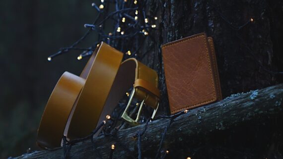 Photo is taken in the darkening forest where the belt and a card wallet is set onto the fallen tree. The whole scene surrounding the yellow leather goods is a bit blue toned. Some small sparkling lights in the background and foreground give it a nice warmer feeling.