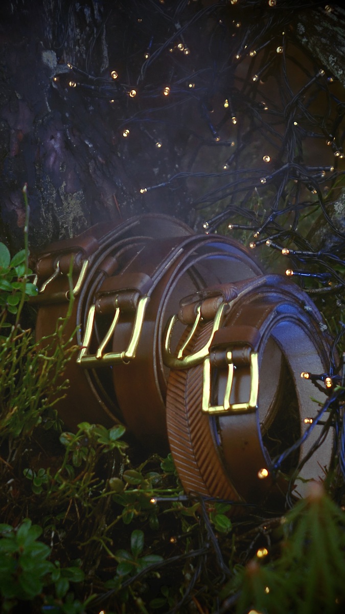 Belts are set in a row towards the camera, leaning to the tree trunk on the forest floor. Leather goods are surrounded by forest plants and there are some decorative small sparkling lights on the top and right part of the photo.