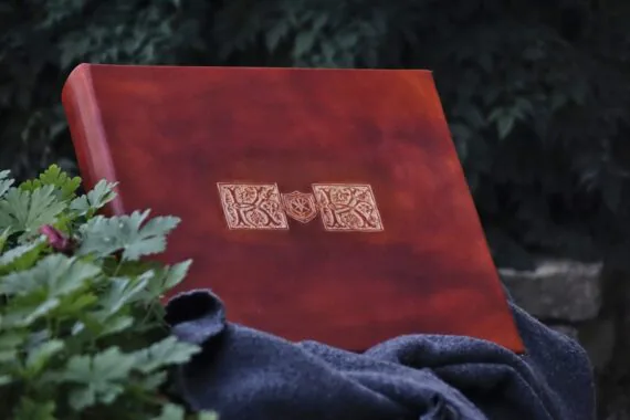 Square shaped album is placed onto the woolen blanket on the stone wall. Album is surrounded by rather darker green bushes.
