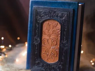 Book has new blue leather covers. The decoration on the front cover is layered, built from multiple layers and parts. The centrepiece is a hand carved image of lilies. This part is not coloured and remains light. The centre is in turn surrounded by next layer of decorated  leather frame.
