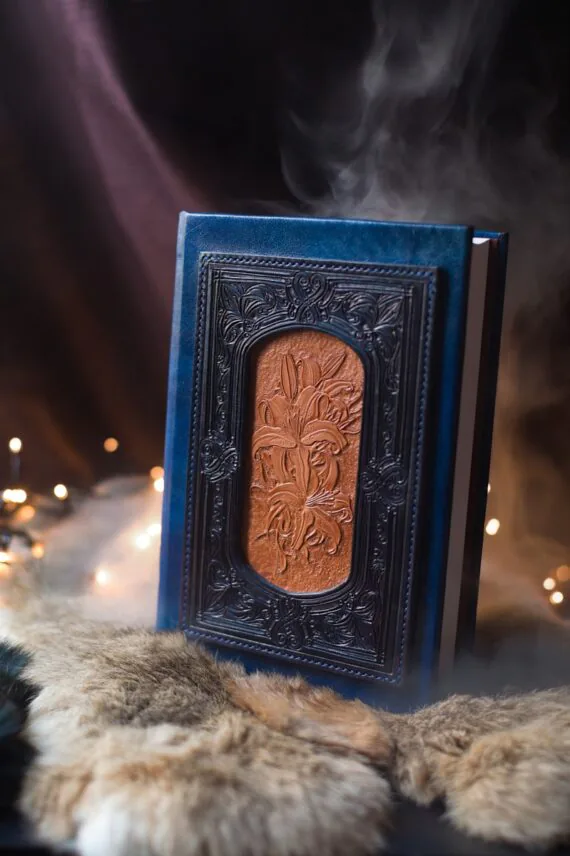Book has new blue leather covers. The decoration on the front cover is layered, built from multiple layers and parts. The centrepiece is a hand carved image of lilies. This part is not coloured and remains light. The centre is in turn surrounded by next layer of decorated  leather frame.