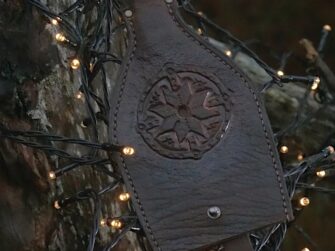 This is a photo of a leather key case in dark brown. Case is decorated by hand carved eight pointed star and ring cross. Key case is surrounded by small electric lights which in torn are hung onto the tree trunk.
