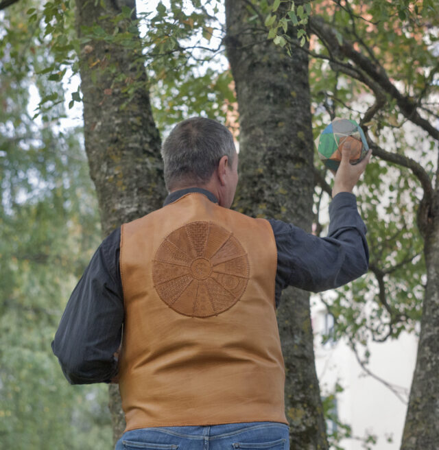 Pildil on vest tellija seljas, kes seisab vaataja poole seljaga. Foto on tehtud õues kenade lehtes kaskede all.