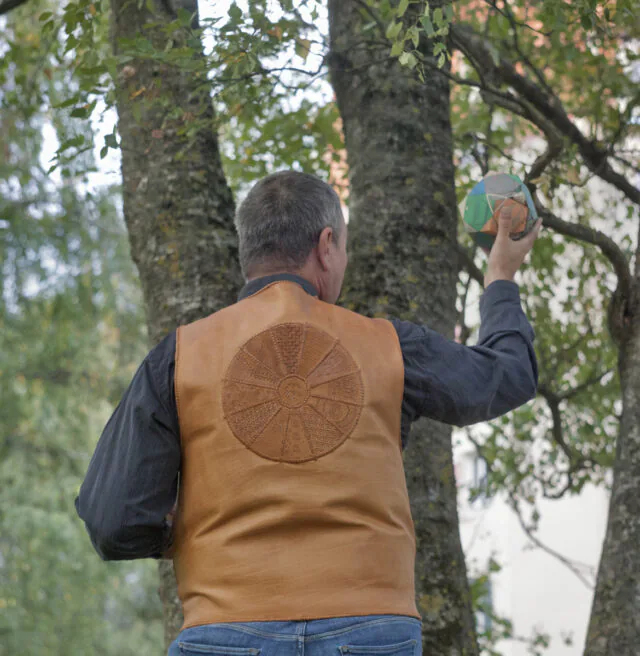 Pildil on vest tellija seljas, kes seisab vaataja poole seljaga. Foto on tehtud õues kenade lehtes kaskede all.